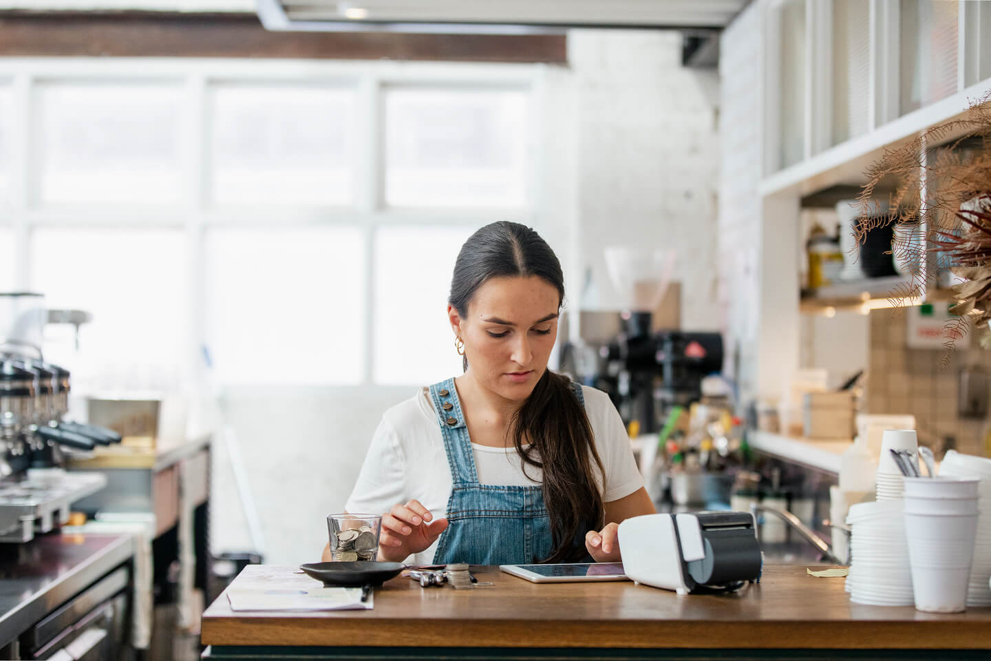 A woman in a café, wearing denim overalls, focuses on a tablet. She's surrounded by coffee-making equipment, creating a busy yet calm atmosphere.