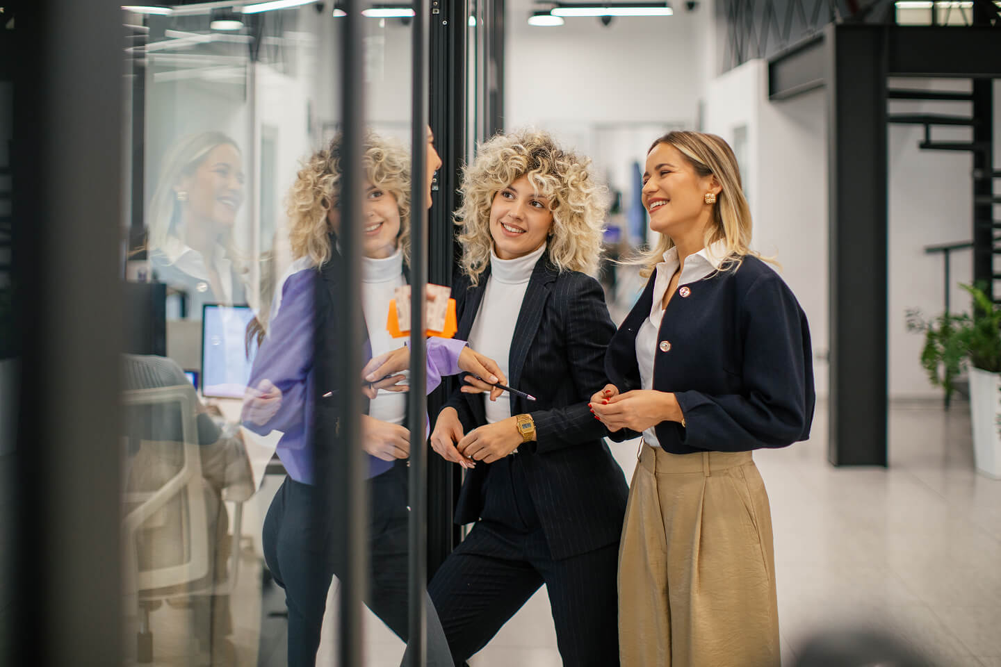 Two women in business attire stand smiling and talking in a modern office hallway, with their reflections visible in the glass wall beside them.