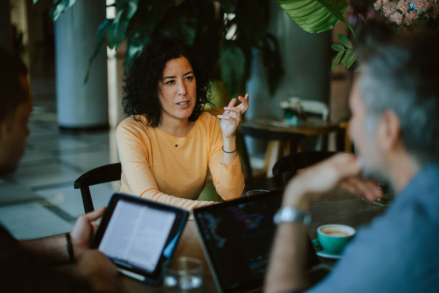 A woman in a yellow sweater engages in a discussion with two people at a table, one with a tablet. The setting is a café with a relaxed atmosphere.