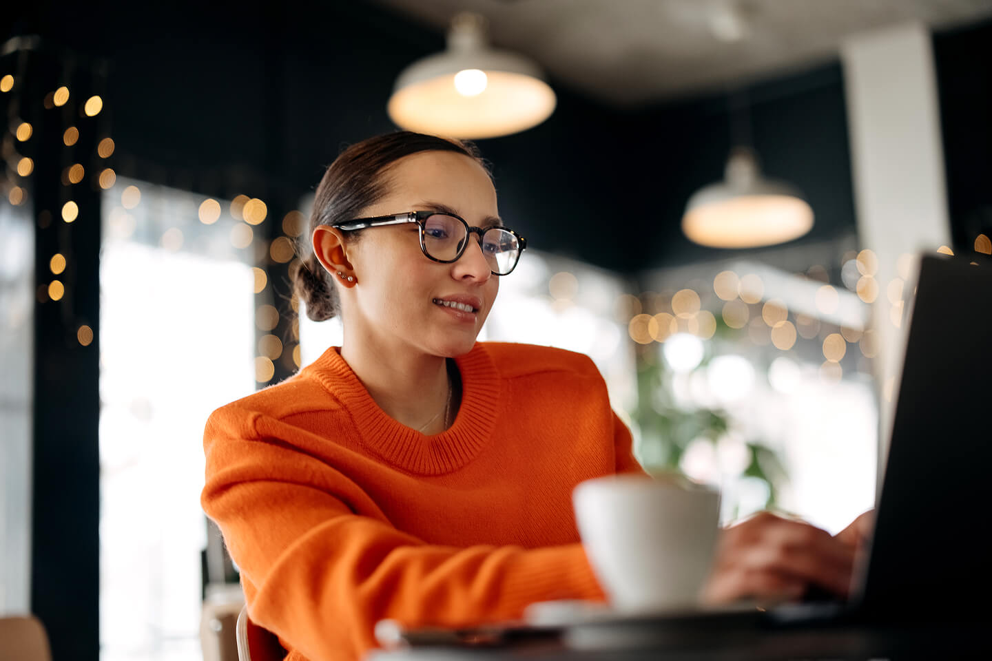 A person in an orange sweater works on a laptop in a cozy café with warm lighting and blurred fairy lights in the background, creating a focused and inviting atmosphere.