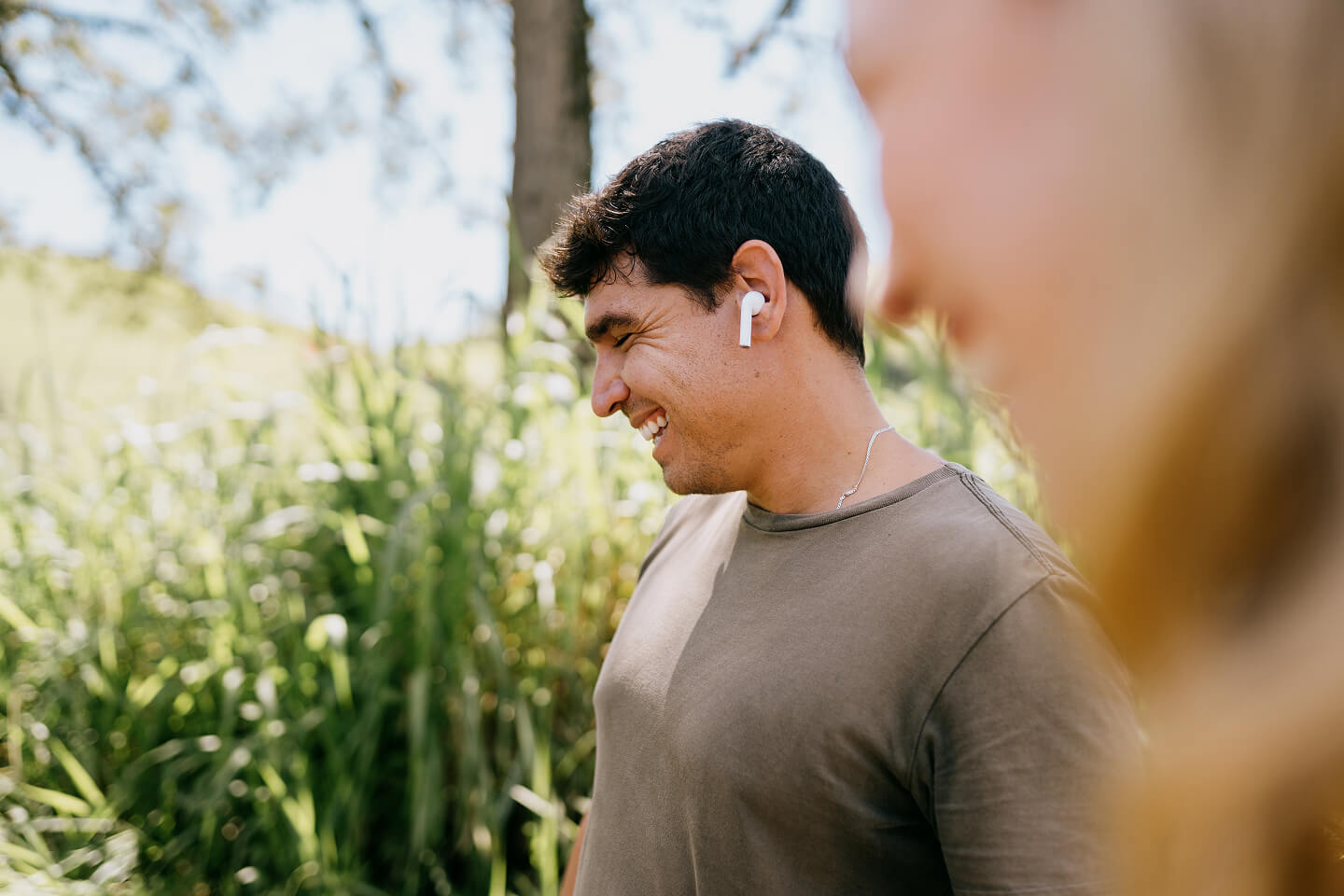 A man wearing wireless earbuds smiles while walking outdoors on a sunny day. He is surrounded by lush greenery, creating a relaxed, joyful atmosphere.